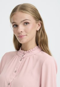 Light pink blouse with a ruffled collar, buttoned front, and soft fabric. The sleeves are slightly gathered at the shoulders.