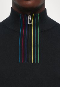 Dettaglio di un maglione nero a maglia con zip frontale e righe verticali in blu, rosso, viola, giallo, turchese e verde lungo la zip.