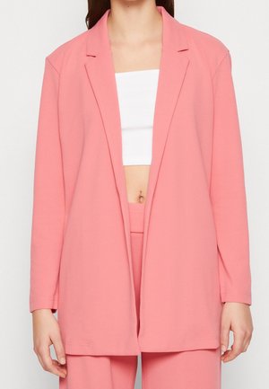 Kvinde iført en løs lyserød blazer og matchende bukser over en hvid crop top, der viser en lille navlepiercing.
