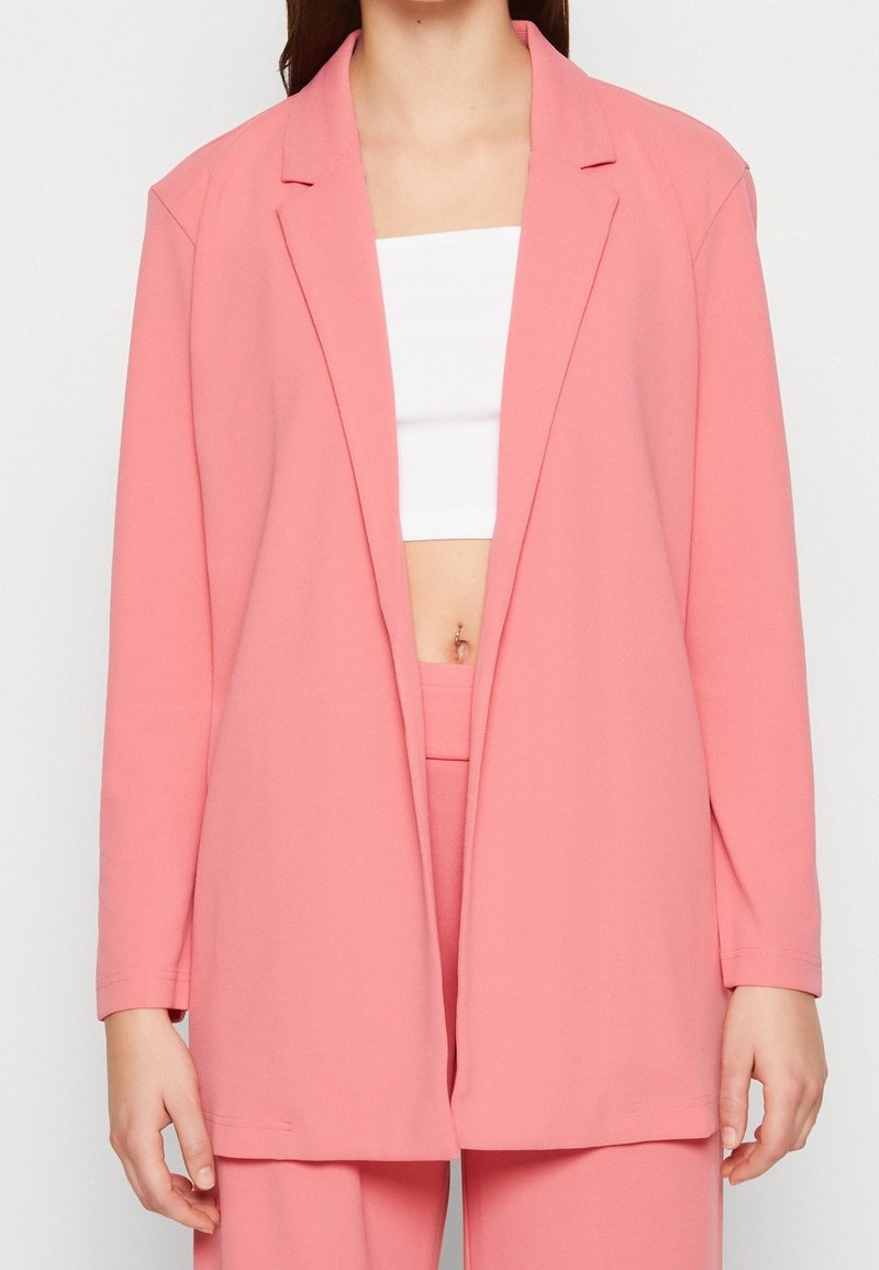 Femme portant un blazer rose ample et un pantalon assorti par-dessus un crop top blanc, montrant un petit anneau au nombril.