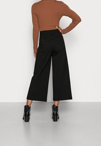 Mustat leveät housut, jotka on valmistettu tekstuurikankaasta, yhdistettyinä tiukkaan ruskeaan ribattuun puseroon ja mustiin korkokenkiin.