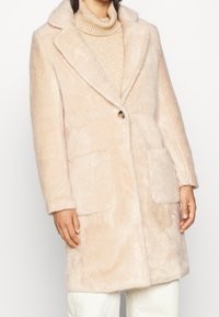 Abrigo de un beige claro, hecho de un material suave y suave. Cuenta con un cuello en pico, cierre de botón único y dos bolsillos delanteros.
