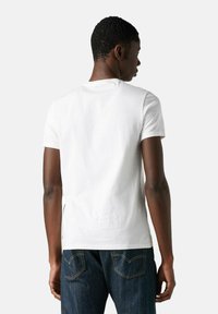 Mand iført en ensfarvet hvid kortærmet t-shirt og mørkeblå jeans, vendt væk fra en lys grå baggrund.