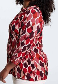 Donna che indossa una camicetta a maniche lunghe con un motivo ovale sovrapposto rosso, rosa e nero e pantaloni bianchi, vista di lato.