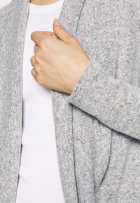 Jasnoszary dzianinowy kardigan o swobodnym kroju, z teksturowanej tkaniny, z otwartym przodem i długimi rękawami, noszony na białej prążkowanej bluzce.