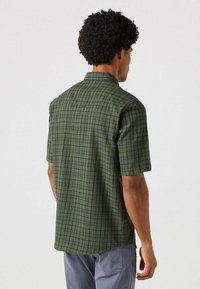 Chemise à manches courtes de couleur vert foncé avec un motif à carreaux bleus, dotée d'un col et d'une ourlet droit, fabriquée à partir d'un tissu texturé.