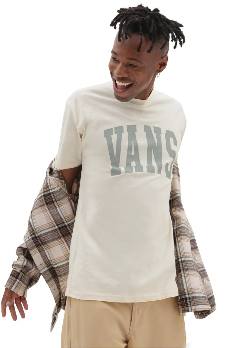 Camiseta crema con "VANS" en grandes letras grises, llevada debajo de una camisa de franela a cuadros beige y marrón, combinada con pantalones beige claros.