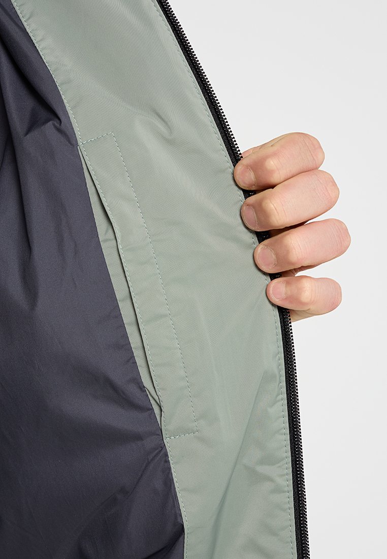 Die Innenseite der leichten Jacke zeigt ein grünes Futter mit einer glatten Oberfläche. Ein schwarzer Reißverschluss und eine kleine Tasche auf der linken Seite runden das Design ab.