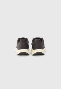 Une paire de chaussures de sport noires avec des semelles blanches vue de l'arrière sur un fond gris clair uni.