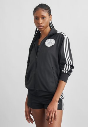 Γυναίκα με πλεγμένα μαλλιά που φοράει μαύρο μπουφάν Adidas και σορτς με λευκές ρίγες, στέκεται μπροστά σε απλό γκρι φόντο.