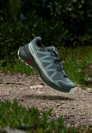 Πράσινο και γκρι παπούτσι τρεξίματος μονοπατιού Salomon με Gore-Tex, αιωρούμενο πάνω από έναν χαλικόδρομο σε δασικό περιβάλλον.