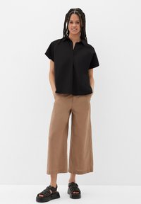 Zwarte korte mouwen shirt met een kraag, gecombineerd met lichtbruine wijde broek en zwarte sandalen met bandjes. Mini-plooi-detail aan de voorkant van het shirt.