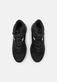 Schwarze Sportschuhe mit einem Mesh-Obermaterial, strukturiertem Design, reflektierenden Akzenten und schwarz-weißen gemusterten Schnürsenkeln. Sichtbare dämpfende Sohle.