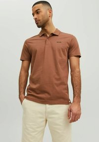 Hnedé polo tričko s krátkymi rukávmi, golierom na dva gombíky a malým čiernym logom "R.D.D." na ľavej hrudi. Mäkký bavlnený materiál.