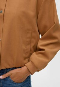 Veste bomber beige en tissu lisse, avec fermeture éclair sur le devant, poignets côtelés et poche latérale, présentée avec une main dans la poche.