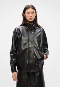 Crna kožna jakna s visokim ovratnikom, kontrastnim šavom, dva bočna džepa i ležernim krojem, s kopčanjem na dugme sprijeda.