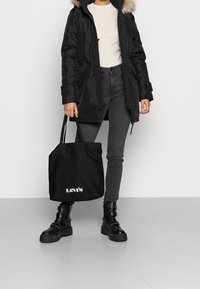 Černá parka s umělou kožešinou na kapuci, šedý svetr, tmavé džíny a robustní černé boty. Černá taška přes rameno s bílým logem "Levi's".