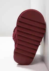 Suela de zapato burdeos con una superficie texturizada, que presenta surcos horizontales para mayor agarre y una forma redondeada. Bordes de tela suave visibles.