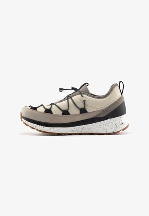 Zapatilla beige y negra con un upper texturizado, cordones elásticos, una suela blanca moteada y una forma curva para mayor comodidad y soporte.