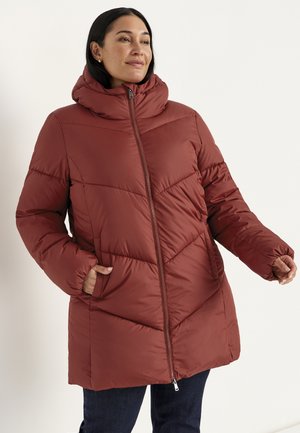 Woman wearing a rust-colored puffer jacket with a high collar, zip closure, and hands in side pockets, standing against a plain light background.