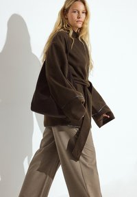 Braune Fleece-Jacke im Wickelstil mit weiten Ärmeln und passendem Gürtel, kombiniert mit hellbraunen, weit geschnittenen Hosen. Weiche Textur und lässige Passform.