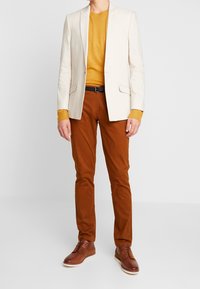 Ljus beige kavaj över en senapsgul tröja, som är sparat med bruna byxor och bruna skor. Kavajen har två framfickor och en modern passform.