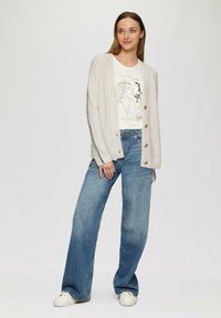 Jasnobeżowy dziergany kardigan zapięty na guziki, nałożony na kremowy t-shirt z grafiką. Do tego szerokie niebieskie dżinsy o luźnych nogawkach i białe trampki.
