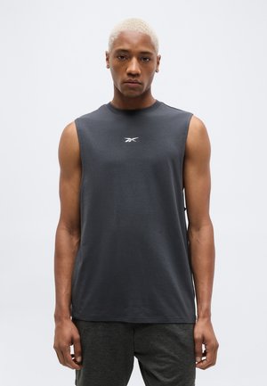 Άνδρας με κοντά ξανθά μαλλιά φορώντας αμάνικο σκούρο γκρι μπλουζάκι Reebok και αντίστοιχα σκούρα γκρι σορτς, στέκεται μπροστά σε απλό ανοιχτό φόντο.