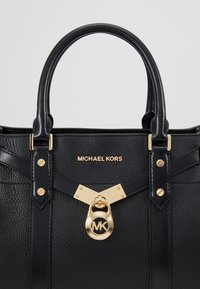 Sac à main Michael Kors en cuir noir avec des ferrures dorées, des poignées rembourrées et un verrou rond doré avec le logo MK à l'avant.