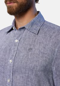 Camicia in lino azzurro chiaro con colletto a bottoni, bottoni bianchi e un piccolo logo. Presenta un tessuto testurizzato con un design casual.
