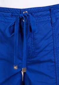 Närbild av ljusblå byxor med tygdragsko knuten i en rosett med metalldetaljer och silverfärgad knappstängning i midjan.