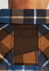 Zbliżenie na niebiesko-brązową tkaninę w kratę z czarną łatą z prostym haftowanym wzorem na ubraniu.