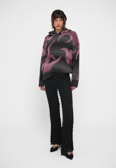 Πλεκτή ζακέτα με βαθμιαία γκρι και ροζ αφηρημένο σχέδιο, συνδυασμένη με μαύρο υφασμάτινο παντελόνι που φουσκώνει, με ραβδώσεις και μυτερά παπούτσια.