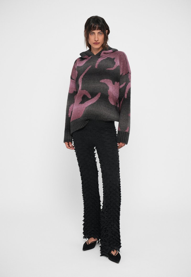 Hoodie in maglia con un motivo astratto in gradiente grigio e rosa, abbinato a pantaloni neri a campana con texture e dettagli arricciati, indossati con scarpe a punta.