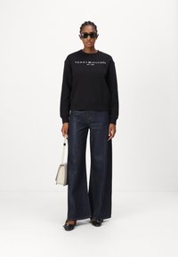 Sudadera negra con logo blanco, combinada con jeans de mezclilla oscuros de pierna ancha. Zapatos negros y un bolso crema completan el look.