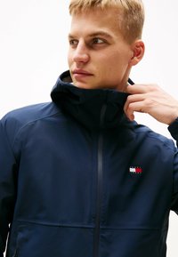 Chaqueta con capucha de color azul marino, hecha de tejido ligero, que cuenta con un cierre delantero, un parche con el logo en el pecho y una textura suave.