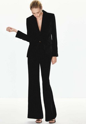 Une femme porte un costume noir sur mesure avec un pantalon évasé et des sandales sur un fond blanc uni.