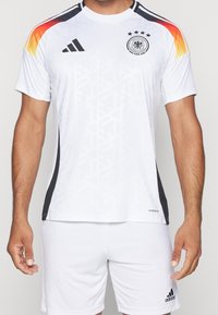 Man som bär en vit tysk fotbollströja med svarta logotyper och gradiente orange-röda ränder på axlarna, tillsammans med vita Adidas-shorts.