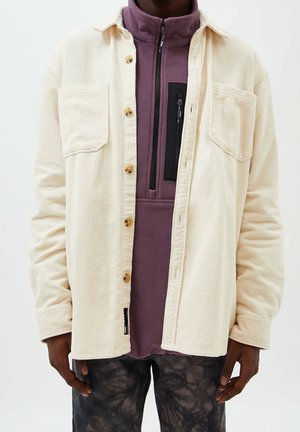 Mann trägt cremefarbenes Cordhemd mit Knöpfen über einem violetten Fleece-Pullover mit schwarzer Reißverschlusstasche und dunklen Batikhosen, Hände entspannt an den Seiten.