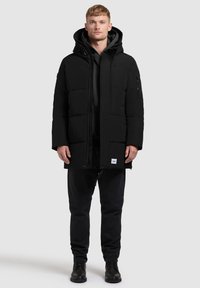 Svart puffjacka med huva, med dragkedja, sidofickor och texturerad yta. Kombineras med svarta byxor och stövlar.