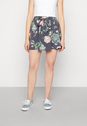 Mujer vistiendo una falda azul marino con estampado floral, cinturón atado a la cintura, top blanco sin mangas con botones y zapatillas grises con cordones blancos.