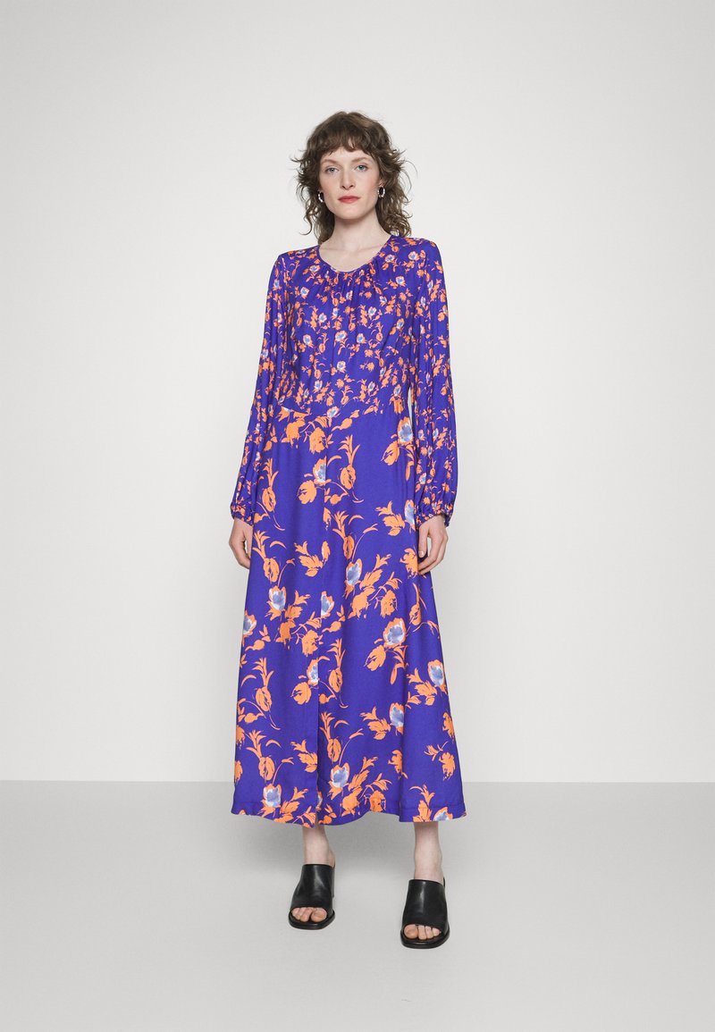 Vestido de mangas compridas, até ao chão, em um vibrante roxo com padrões florais em laranja e azul. Tecido suave com detalhe de decote franzido.