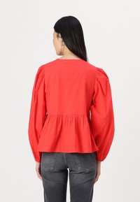 Top peplum rosso con maniche lunghe a sbuffo e vita arricciata, realizzato in tessuto leggero. Jeans grigi visibili sul fondo.