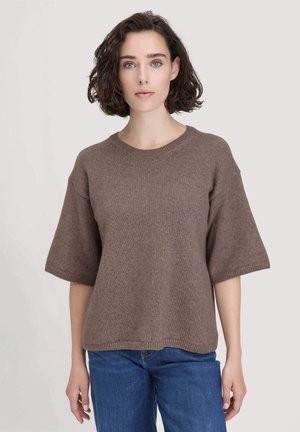 Frau mit kurzen, lockigen, dunklen Haaren, die einen lockeren, braunen Strickpullover mit kurzen Ärmeln und blaue Jeans trägt, vor einem einfarbigen, hellen Hintergrund.