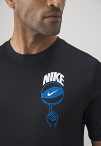 Mann trägt ein schwarzes Nike-T-Shirt mit einem blauen Basketball-Graphic, der auf einem Finger gedreht wird, und dem Text "NIKE" über dem Ball.