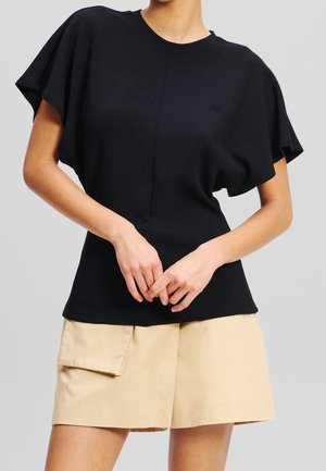 Persoon draagt een zwart kortermig shirt met brede mouwen en beige korte broek met een klepzak aan de zijkant.