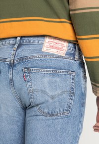 Hellblaue Jeans mit geradem Schnitt, sichtbaren Nähten und einem roten Levi's-Tag an der Rückentasche. Getragen mit einem grün-gelb gestreiften Hemd.