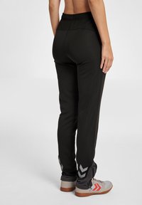 Schwarze Sporthose mit elastischem Taillenbund, ausgestattet mit grauen Chevron-Mustern am Saum und aus strukturiertem Stoff. Getragen mit neutralen Sportschuhen.