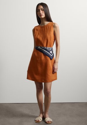 Donna in piedi con abito arancione senza maniche, sciarpa blu navy con fantasia annodata in vita e sandali beige su sfondo semplice.