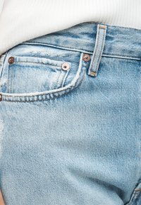 Primer plano del bolsillo de unos vaqueros de denim azul claro con remaches de cobre y camisa blanca metida por encima de la cinturilla.
